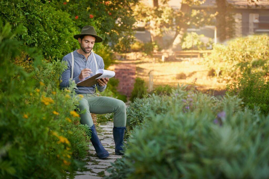 Mann, Gartenarbeit und Klemmbrett für Outdoor-Wachstum, Planung und Forschung von Pflanzen oder Blumen. Junge Arbeiter oder Gärtner mit Checkliste im Hinterhof für kleine Unternehmen, Landwirtschaft und Inspektion Notizen 