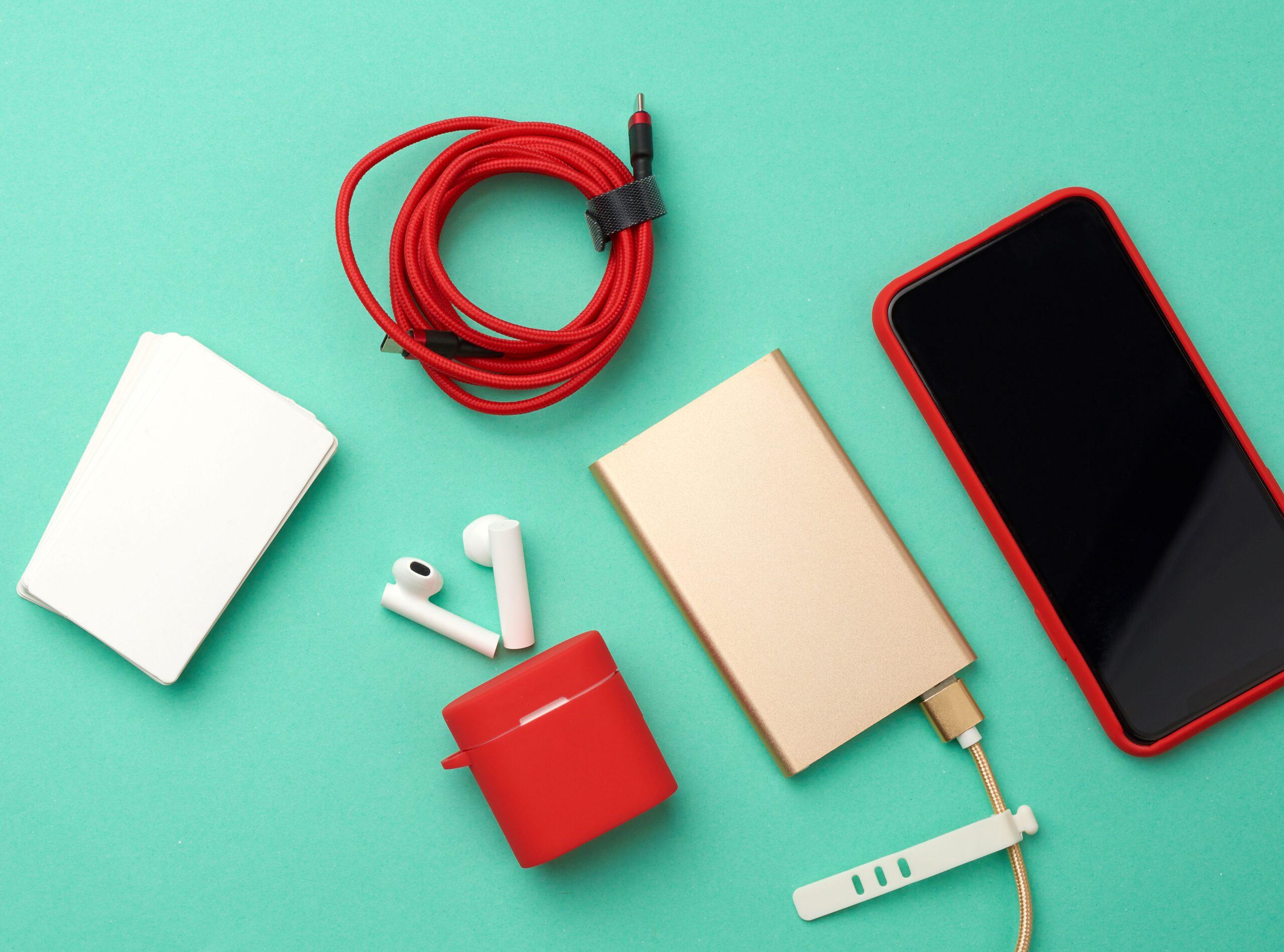 goldene Powerbank mit Kabel, rotes Smartphone mit leerem schwarzen Bildschirm, Box mit Kopfhörern, leere Visitenkarte aus Papier