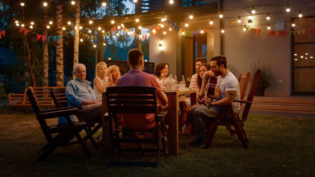 Am Esstisch sitzend, spielt ein hübscher junger Mann Gitarre für einen Freund. Familie und Freunde hören Musik auf dem Sommerabend Gartenparty Feier.